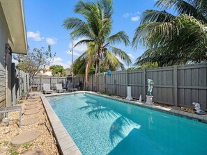 A rectangular swimming pool with a wooden deck on one side, surrounded by a wooden fence, palm trees, and a clear blue sky. - Cozy Haven Oasis: Pool| Los Olas |Wilton Manors (Wilton Manors)