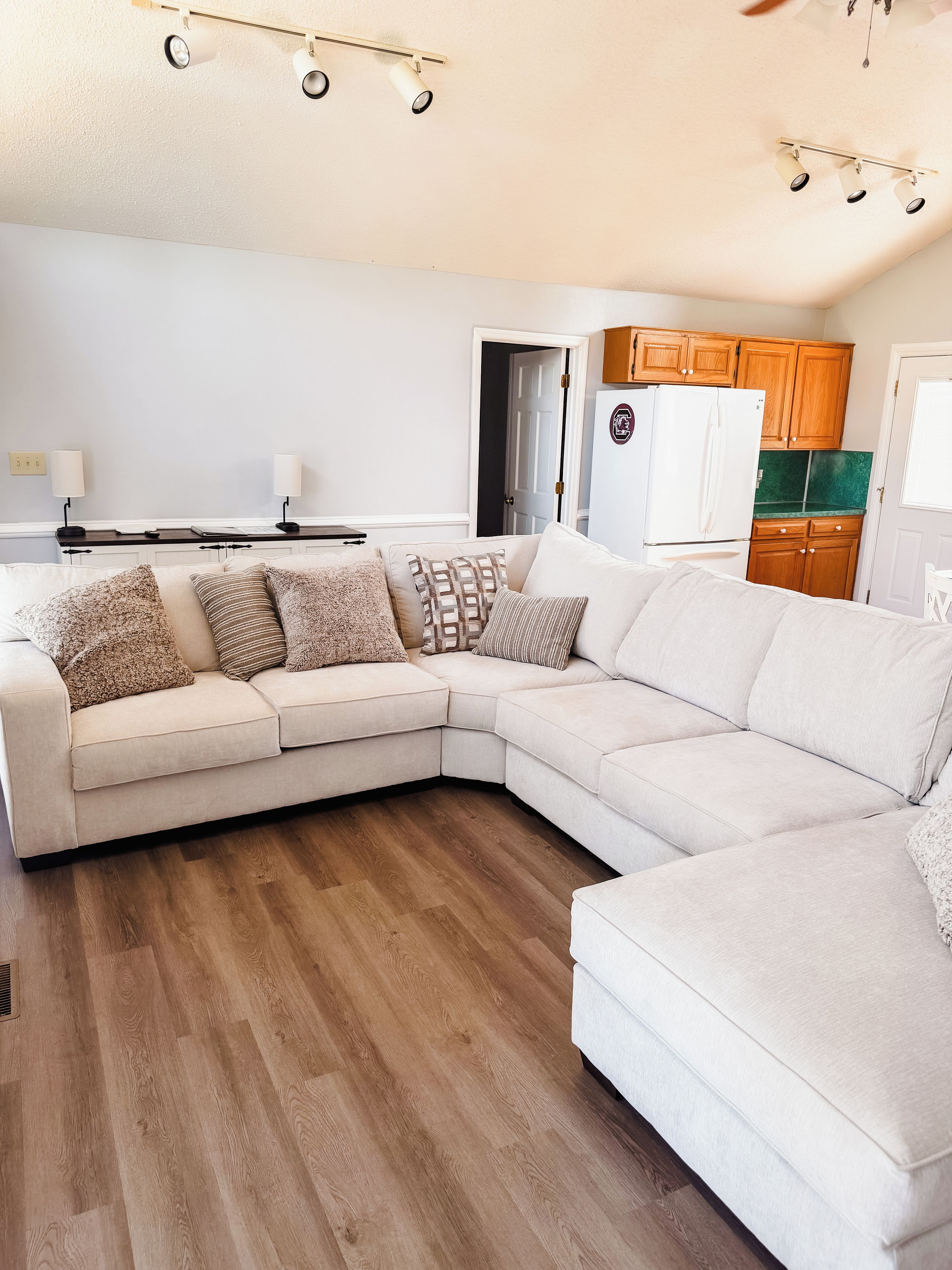Un salon moderne avec un canapé d’angle, un plancher en bois et un coin cuisine à l’arrière-plan.