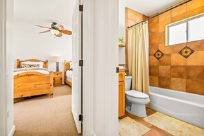 A bedroom with a wooden bed, a nightstand, a bedside table, a ceiling fan, and a bathroom with a bathtub, toilet, and shower curtain. - Great Reviews, Large Rooftop Deck W/Parking, Stunning (San Diego)