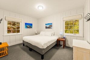 Ein Schlafzimmer mit einem Bett, zwei Fenstern mit Jalousien, einem Nachttisch mit Lampe und einem kleinen Holzhocker.