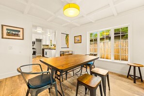 A dining area with a wooden table and chairs, a kitchen in the background, and a large window with a view of a wooden fence.