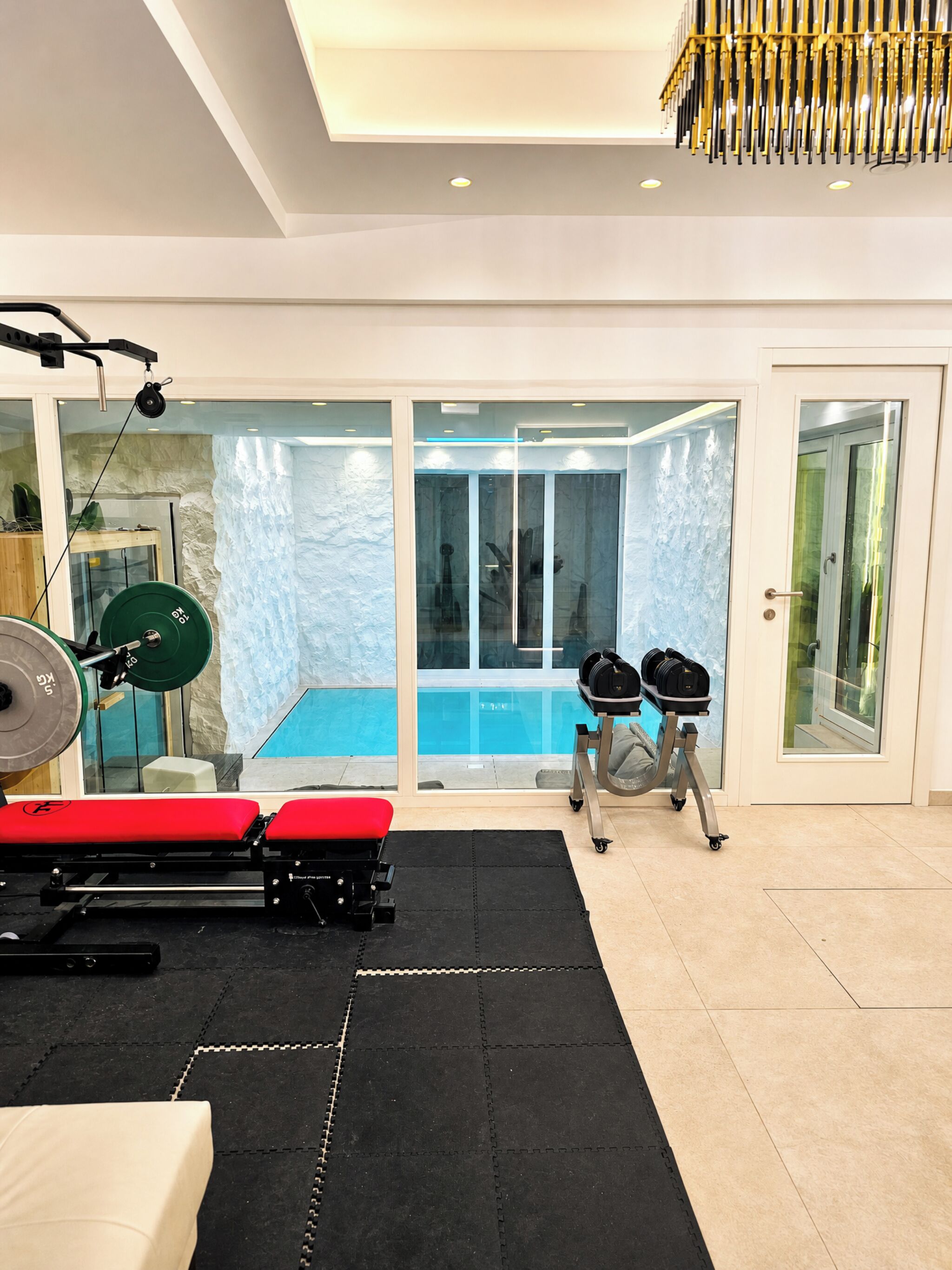 Ein Heimfitnessraum mit Langhantelbank, Kniebeugestange und Gewichtsplatten mit Blick auf einen Swimmingpool.
