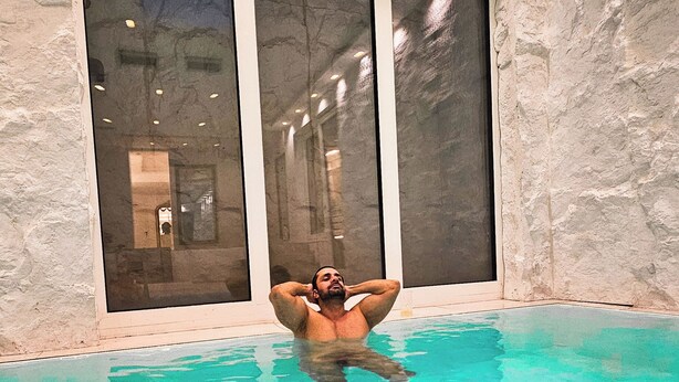 A man relaxing in a clear blue indoor pool with stone walls and large windows.