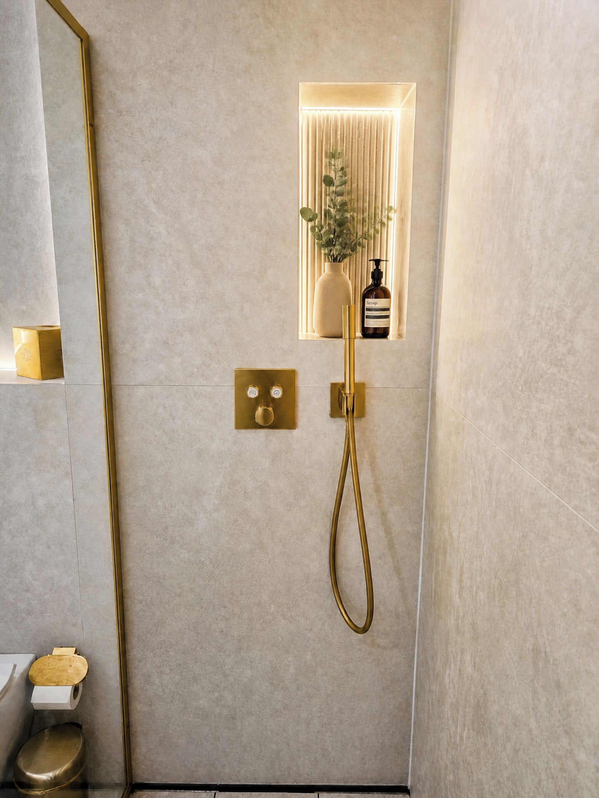 A modern bathroom with a gold shower head, a wall-mounted control panel, and a small window with a plant.