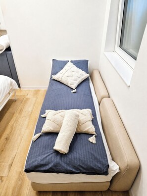 A single bed with a dark blue mattress and two pillows, a window with blinds, and a wooden floor. - Neubau City Wohnung (Pool+Gym+Sauna extra buchbar) (München)