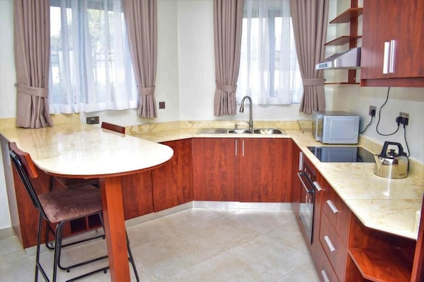 A kitchen with a marble countertop, wooden cabinets, a sink, a microwave, and a kettle.