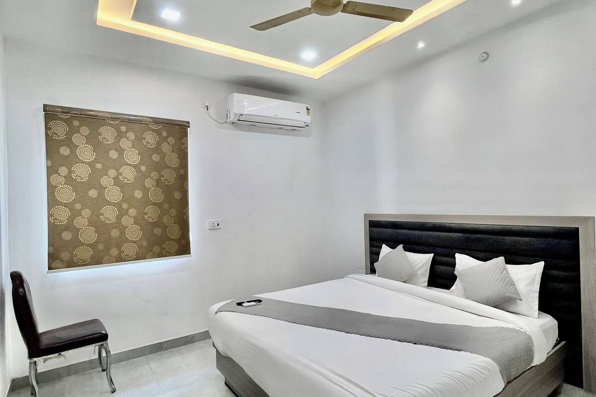A modern bedroom with a bed, a ceiling fan, an air conditioning unit, a patterned window treatment, and a chair.