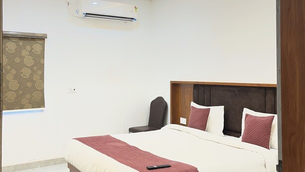 A modern bedroom with a bed, ceiling fan, air conditioning unit, and a window with a patterned curtain. - Hotel Girraj Stay Inn  (Mathura)