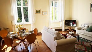 En stue med sofa, fjernsyn, spisebord med stole og to vinduer med gardiner.