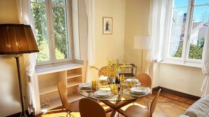 Spiseplads med et rundt bord, stole, en lampe og udsigt til grøn bevoksning.