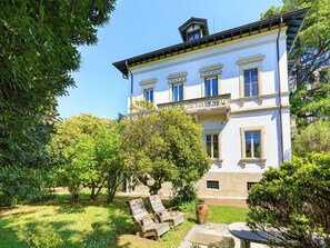 Un grand bâtiment blanc, doté de nombreuses fenêtres et d’un balcon, entouré d’une végétation luxuriante et d’arbres.