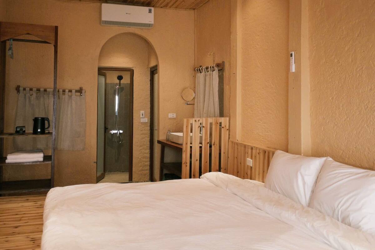 Une pièce avec un plafond en bois, un lit avec du linge de lit blanc, un banc en bois, une petite table avec une tasse et un espace douche.