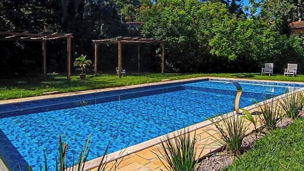 Una piscina rectangular con borde de azulejos, rodeada de césped y árboles.