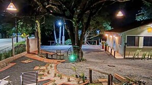 Vista nocturna de una propiedad con un gran árbol, asientos al aire libre y un sendero bien iluminado.