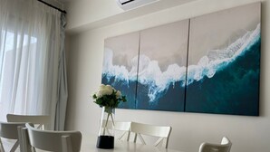 Ein Essbereich mit einem weißen Tisch, weißen Stühlen, einer Vase mit Blumen und einem großen Gemälde einer Meereswelle an der Wand.