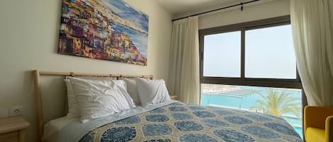 A bedroom with a bed, a painting, a window with a view, and a hanging light fixture.