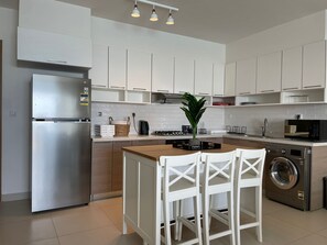 A modern kitchen with white cabinets, a stainless steel refrigerator, a washing machine, and a wooden island with chairs. - Chalet 3 Bedrooms LR - 404 in Marassi (El Alamein)