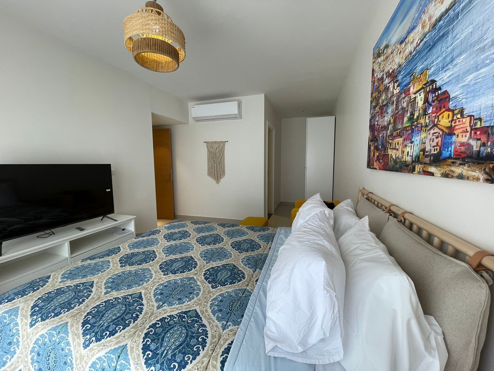 A bedroom with a bed, a television, a macrame wall hanging, and a painting of a coastal town.