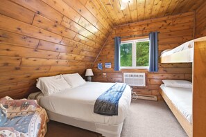 A wooden cabin bedroom with a bunk bed, a single bed, a window with curtains, a wall-mounted heater, and a small table with a lamp. - Juniper Presents: Chalet #74 Walk to Pool, Courts & Resort Amenities (Stevensville)