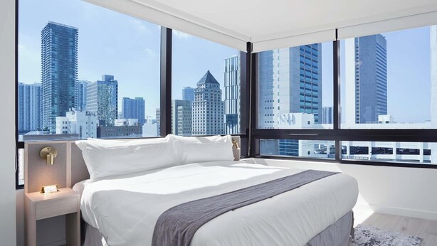 Een moderne hotelkamer met een groot bed, een nachtkastje en uitzicht op een stadshemelbeeld door grote ramen.