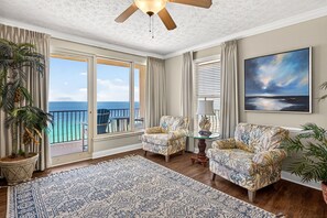 A room with a large window, a ceiling fan, two patterned armchairs, a table with a lamp, and a painting on the wall. - Spacious 3-bedroom condo with a Sunset View in tranquil Panama City Beach (Panama City Beach)