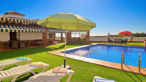 Una piscina con sillones de descanso y sombrillas, rodeada de un césped cuidado y un edificio con tejado de tejas.