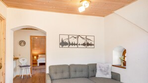 A cozy living room with a grey sectional sofa, a small blue ottoman, a wall-mounted picture of mountains, and a wooden ceiling. - Apartment 'Fewo Edelweiss' with Private Sun Terrace and Wi-Fi (Breitenberg)