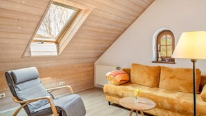 Een gezellig loft slaapkamer met een schuine kap, een houten stoel, een bank, een klein tafeltje en een raam met uitzicht op bomen.