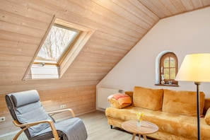 Ein gemütliches Schlafzimmer im Loft-Stil mit schrägem Dach, einem Holzstuhl, einer Couch, einem kleinen Tisch und einem Fenster mit Blick auf Bäume.