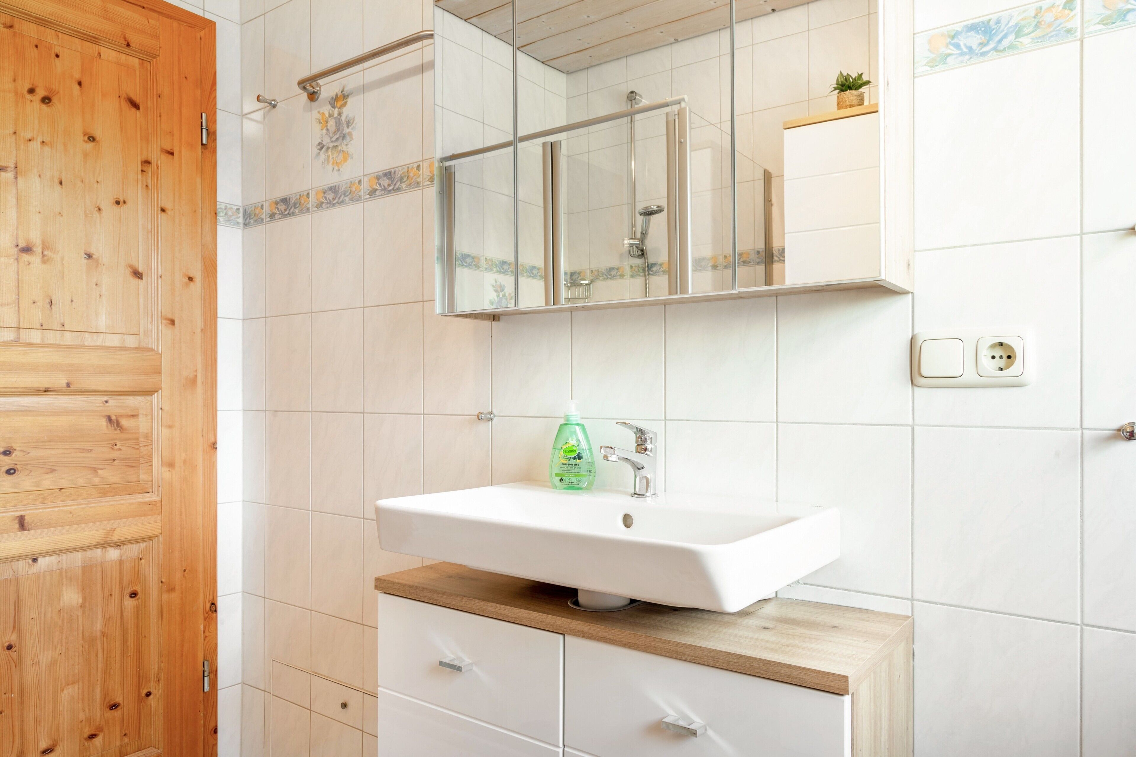 A bathroom with a wooden door, white tiled walls, a white sink with a green bottle, a mirror, and a small plant on the wall.
