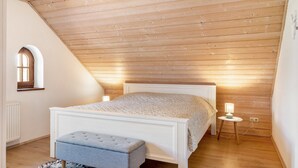 Een slaapkamer met een schuine kap, een bed, een klein tafeltje met een lamp en een raam.