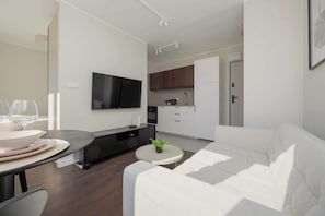 Een moderne woonkamer met een bank, een eettafel met borden en glazen, een flatscreen televisie, een klein rond tafeltje met een plant en een keuken met kasten en een koelkast.