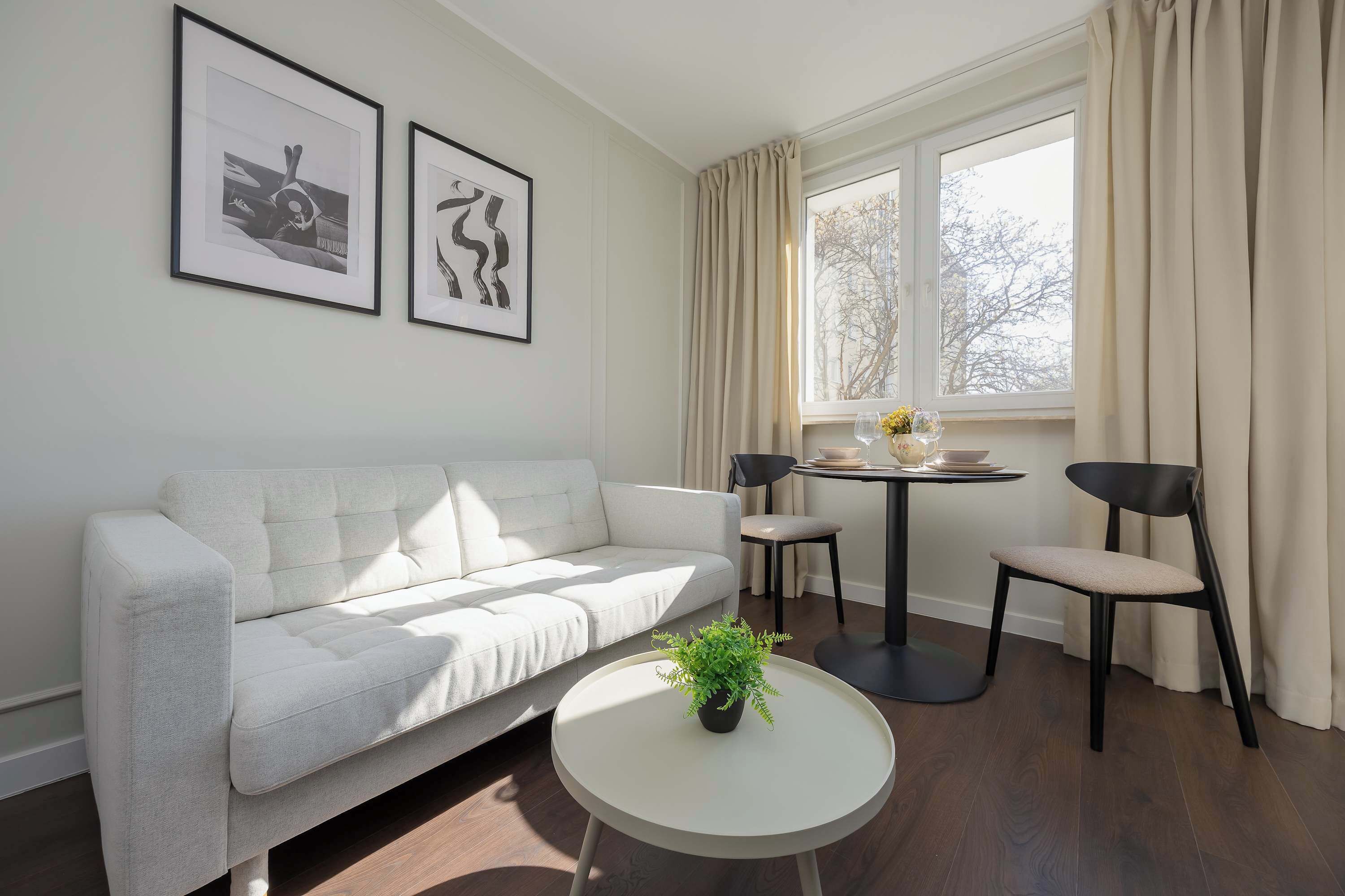 A modern living room with a white sofa, a round coffee table with a plant, two chairs, a small round table with a vase, and framed artwork on the wall.