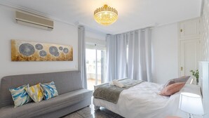 Une chambre moderne avec un canapé gris, un grand lit avec une literie blanche, un lustre et un tableau au mur.
