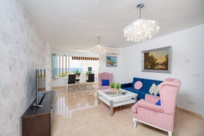 A modern living room with a sofa, armchairs, a coffee table, and a TV. There are two pendant lights, a painting on the wall, and a view through the window. - Fidalsa Bella Vista Junto al mar (El Campello)
