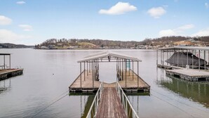 Un molo di legno che conduce a un pontile con una tettoia, acqua calma e una costa lontana con edifici e alberi.