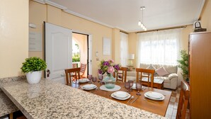 Ett väl upplyst, modernt kök med bänkskivor i granit, en matplats med ett träbord för fyra personer och ett mysigt vardagsrum med en soffa och en lampa.