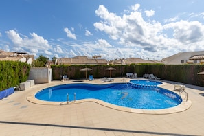 A swimming pool with a curved design, surrounded by lounge chairs and umbrellas, with a view of residential buildings in the background. - Honey Life Villa by Fidalsa (Orihuela)
