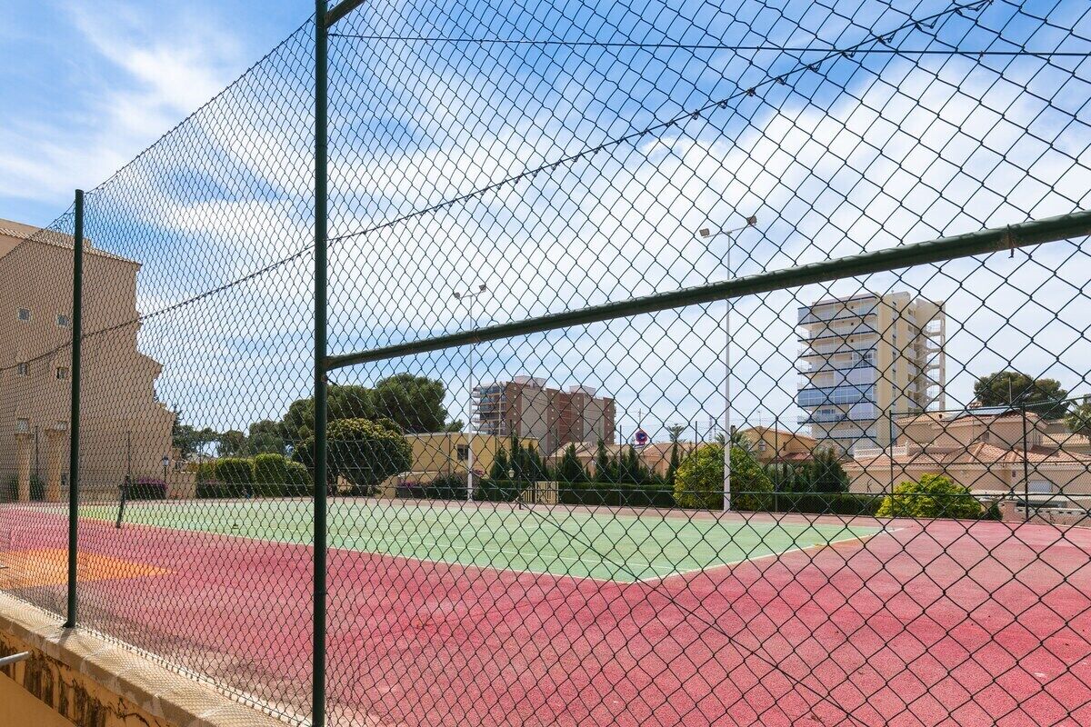 Een omheind tennisterrein met een rode ondergrond, omringd door gebouwen en bomen.