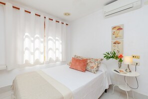 A bedroom with a bed, white curtains, a wall-mounted air conditioner, and a bedside table with a lamp and a vase of flowers. - Fidalsa Feel the Sea (Guardamar del Segura)