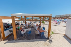 An outdoor terrace with wooden tables and chairs, overlooking a beach with umbrellas and people. - Flamingo Coast by Fidalsa (Orihuela)
