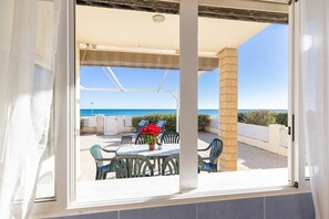 Une vue depuis une fenêtre sur une terrasse extérieure avec une table et des chaises, un pot de fleurs et une vue sur la mer.