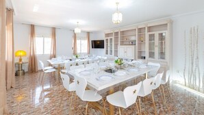A dining area with a large rectangular table set for multiple people, surrounded by white chairs with gold accents. There are two pendant lights above the table, and a television is mounted on the wall. - Fidalsa Grand White House (Mutxamel)