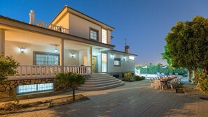 A two-story house with a stone foundation, a balcony, and a paved driveway leading to a dining area with outdoor furniture. - Fidalsa Grand White House (Mutxamel)