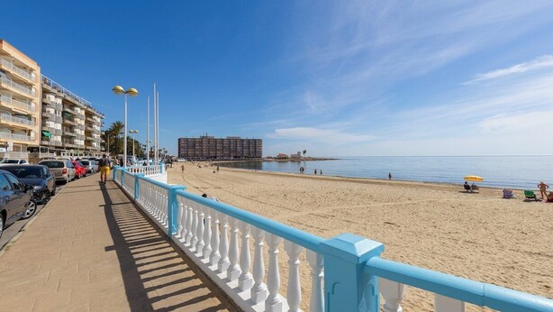 A coastal promenade with a sandy beach, a blue railing, and a row of parked cars.