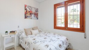 Ein Schlafzimmer mit Bett, Fenster, einem Gemälde, einem kleinen Tisch mit Büchern und einem Regal.