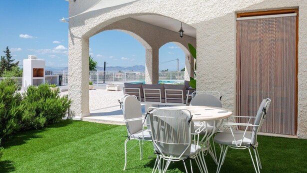 Eine Terrasse mit weißem Tisch und Stühlen, Blick aufs Meer und ein Gebäude mit weißer Fassade.