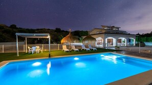 Ein Schwimmbecken mit Liegestühlen, einer überdachten Lounge und einem Gebäude mit Rundbogenfenstern.