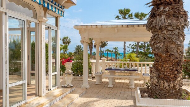 A covered outdoor area with a palm tree, a pergola, and a building with large windows. - Fidalsa Mediterranean Beachfront (Orihuela)
