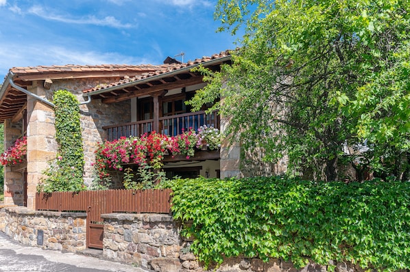 Un edificio de piedra con balcón de madera, rodeado de exuberante vegetación y plantas en flor.